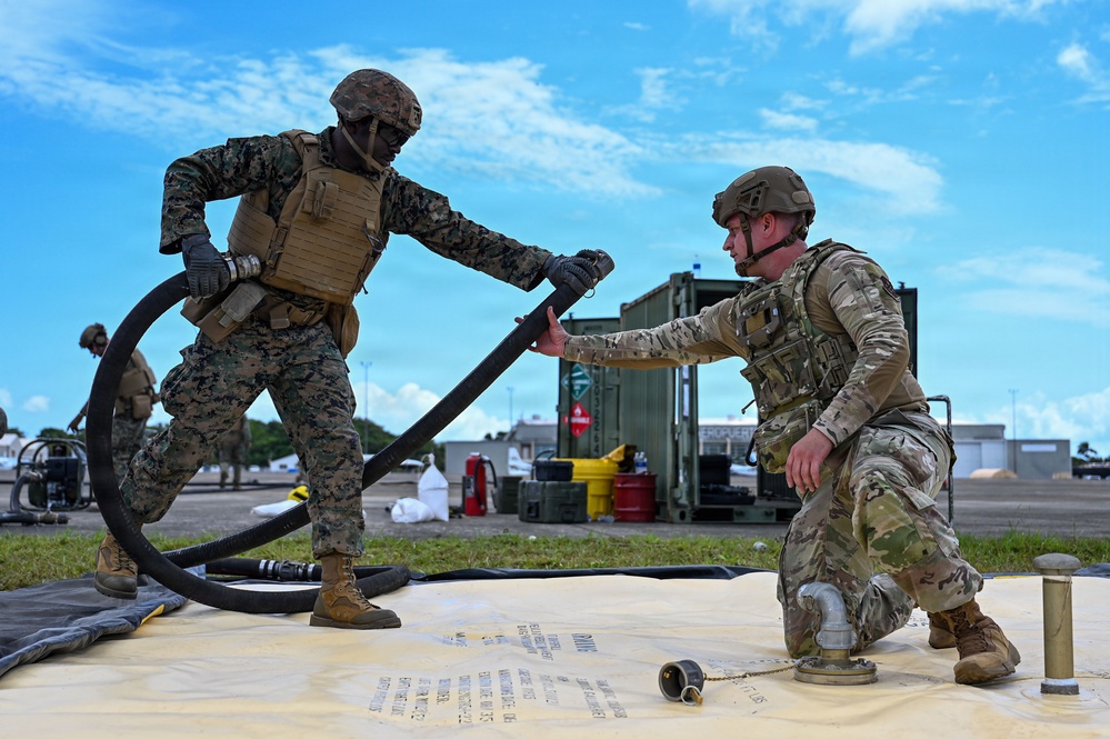 Two military personnel in different camouflage uniforms — one in MARPAT (Marine Corps) and one in OCP (Army/Air Force) — are collaborating on a fuel hose connection exercise on what appears to be a collapsible fuel bladder or berm. They are working with large black flexible hoses at what appears to be a forward area refueling point (FARP) or fuel operations site. The setting is an outdoor military installation or airfield with equipment and containers visible in the background.