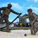 Two military personnel in different camouflage uniforms — one in MARPAT (Marine Corps) and one in OCP (Army/Air Force) — are collaborating on a fuel hose connection exercise on what appears to be a collapsible fuel bladder or berm. They are working with large black flexible hoses at what appears to be a forward area refueling point (FARP) or fuel operations site. The setting is an outdoor military installation or airfield with equipment and containers visible in the background.