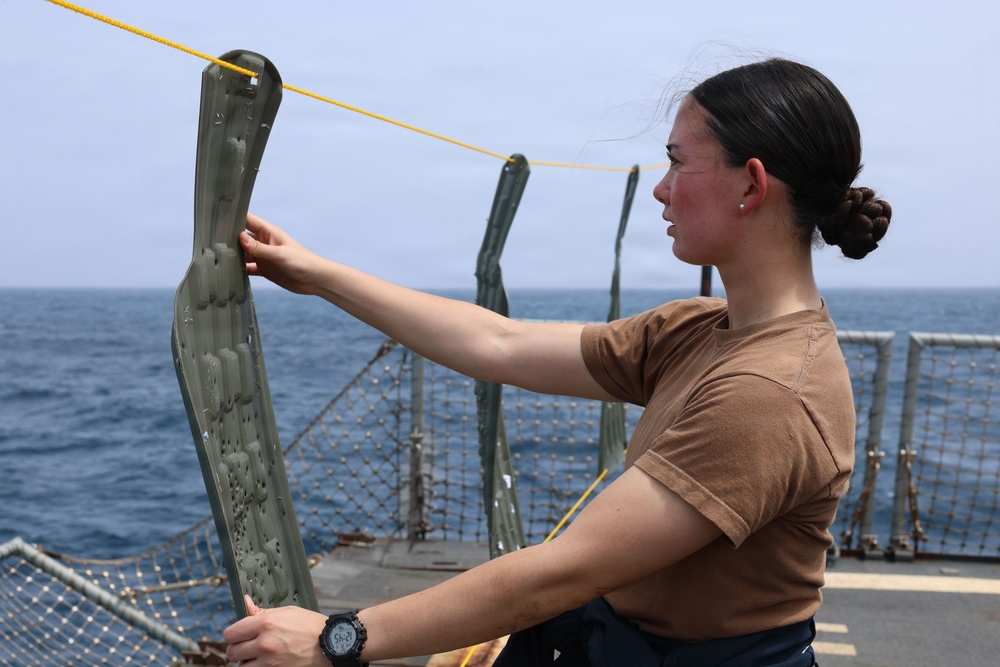 A woman in a tan/brown t-shirt and dark pants is standing on the deck of a naval vessel, holding up and examining a green inflatable item, possibly a life preserver or flotation device. The ship's deck features safety netting, metal stanchions, and a yellow rope line. The ocean and a hazy horizon are visible in the background.