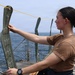 A woman in a tan/brown t-shirt and dark pants is standing on the deck of a naval vessel, holding up and examining a green inflatable item, possibly a life preserver or flotation device. The ship's deck features safety netting, metal stanchions, and a yellow rope line. The ocean and a hazy horizon are visible in the background.