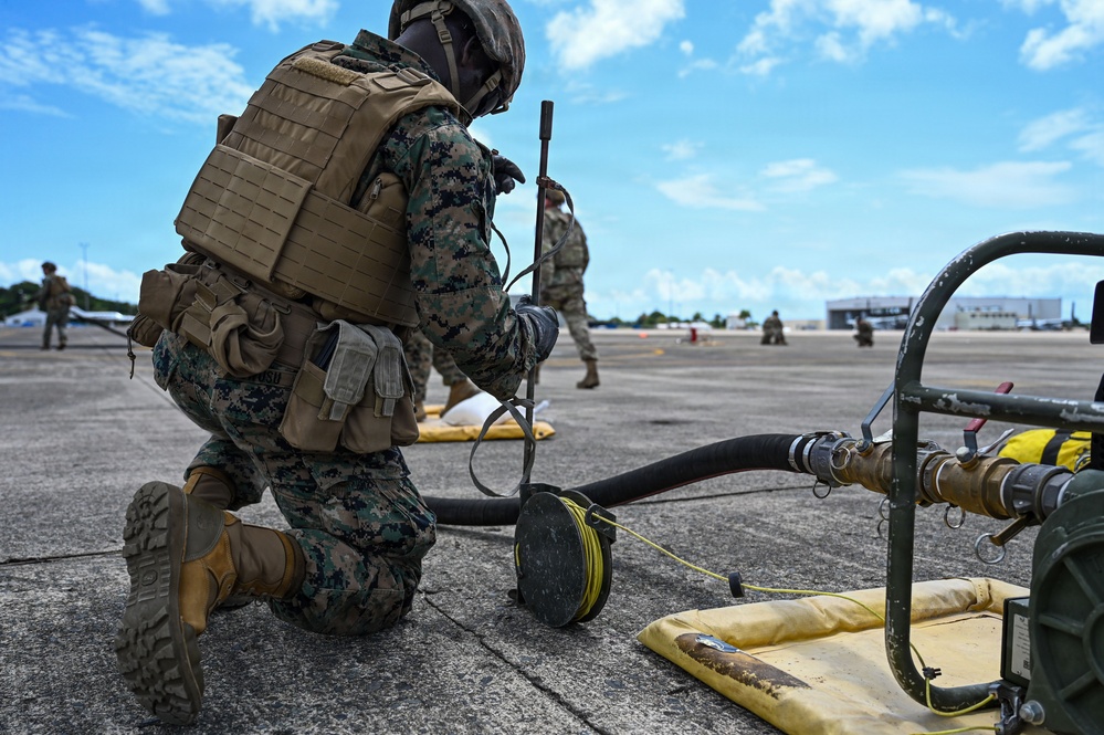 A U.S. Marine in full combat gear and MARPAT camouflage is kneeling on a tarmac, working with fuel hose equipment connected to a refueling apparatus. Additional Marines are visible in the background on what appears to be an airfield or flight line. The scene depicts a forward area refueling point (FARP) or similar tactical fuel operations exercise.