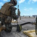A U.S. Marine in full combat gear and MARPAT camouflage is kneeling on a tarmac, working with fuel hose equipment connected to a refueling apparatus. Additional Marines are visible in the background on what appears to be an airfield or flight line. The scene depicts a forward area refueling point (FARP) or similar tactical fuel operations exercise.