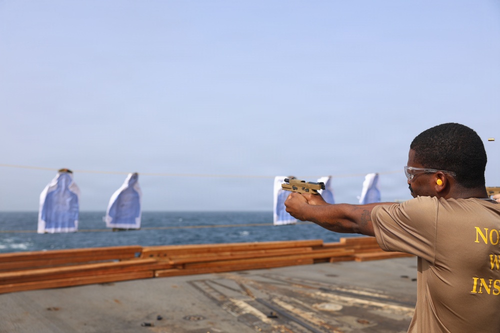 A person is conducting firearms training on what appears to be the deck of a naval vessel. The shooter is aiming a tan/FDE-colored pistol at multiple humanoid paper targets suspended on a line over the water. The individual is wearing proper safety equipment including ear protection and safety glasses.