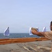 A person is conducting firearms training on what appears to be the deck of a naval vessel. The shooter is aiming a tan/FDE-colored pistol at multiple humanoid paper targets suspended on a line over the water. The individual is wearing proper safety equipment including ear protection and safety glasses.