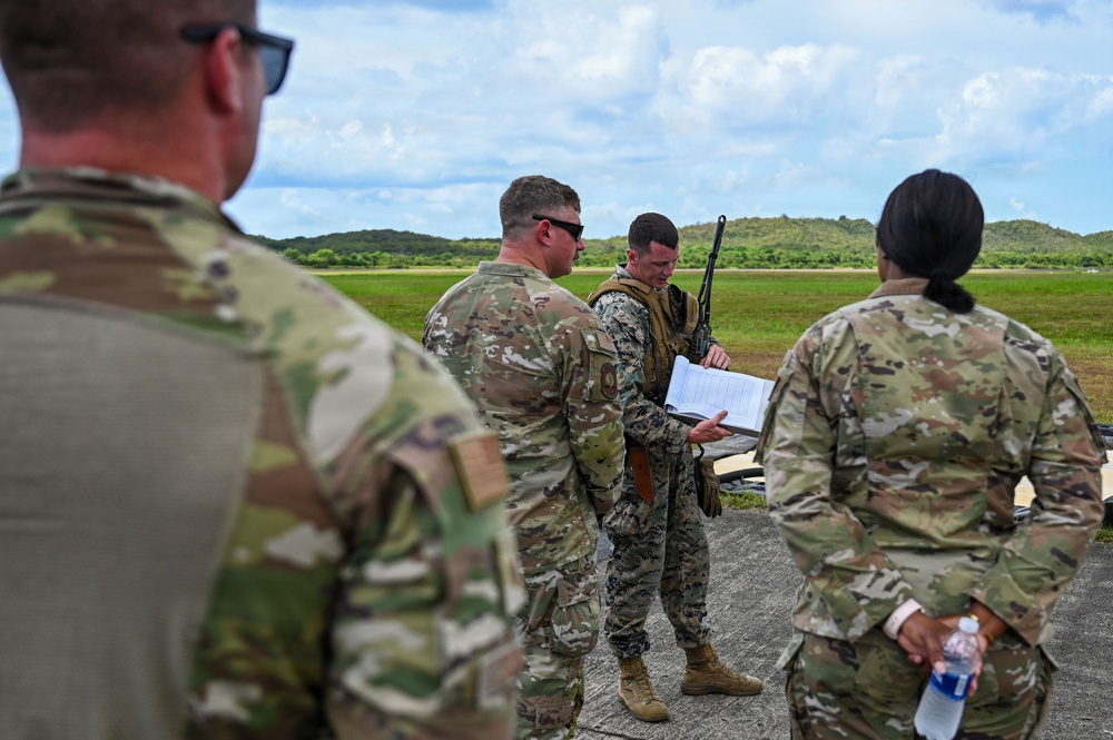 A group of military personnel in different uniforms are gathered outdoors on a paved surface, reviewing documents together. At least two distinct uniform types are visible: US Army OCP (Operational Camouflage Pattern) and US Marine Corps MARPAT digital camouflage. The setting appears to be a training range or airfield with green hills and open fields in the background.