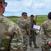 A group of military personnel in different uniforms are gathered outdoors on a paved surface, reviewing documents together. At least two distinct uniform types are visible: US Army OCP (Operational Camouflage Pattern) and US Marine Corps MARPAT digital camouflage. The setting appears to be a training range or airfield with green hills and open fields in the background.