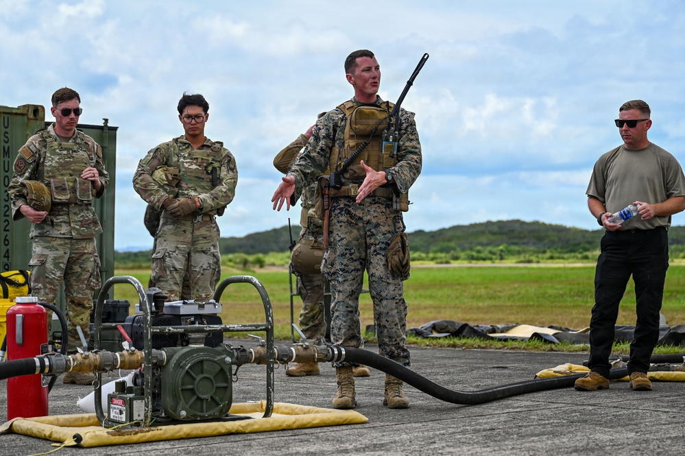 A group of military personnel are gathered outdoors on a tarmac or airfield surface during what appears to be a training or briefing session. A Marine in full combat gear with a rifle is speaking to the group, which includes Air Force personnel in MultiCam uniforms and a civilian or contractor in civilian attire. Fuel or fluid transfer equipment, including a pump and hoses, is visible in the foreground.