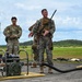 A group of military personnel are gathered outdoors on a tarmac or airfield surface during what appears to be a training or briefing session. A Marine in full combat gear with a rifle is speaking to the group, which includes Air Force personnel in MultiCam uniforms and a civilian or contractor in civilian attire. Fuel or fluid transfer equipment, including a pump and hoses, is visible in the foreground.