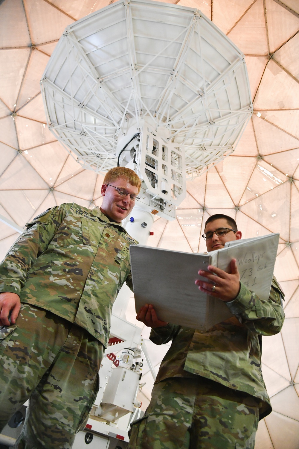 Two military personnel in OCP (Operational Camouflage Pattern) uniforms are reviewing a binder/manual inside a radome facility housing a large satellite dish antenna. The image is taken from a low angle, emphasizing the scale of the parabolic antenna and geodesic dome structure behind them. The soldier on the left has a name tape reading 'VAN GRUBB' visible on their uniform.