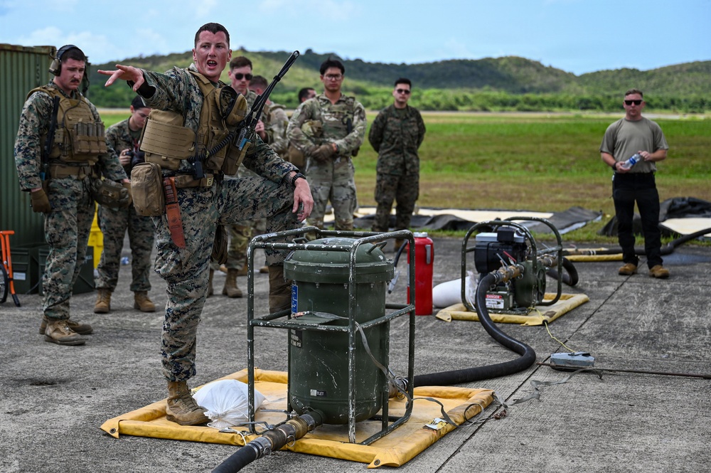 A U.S. Marine in full combat gear and body armor appears to be conducting a briefing or instruction session for a group of military personnel on what appears to be an airfield or tarmac. The instructor is standing on a yellow containment berm next to military fuel handling or pumping equipment, gesturing to direct attention. Multiple service members in various camouflage uniforms are observing the demonstration.