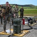 A U.S. Marine in full combat gear and body armor appears to be conducting a briefing or instruction session for a group of military personnel on what appears to be an airfield or tarmac. The instructor is standing on a yellow containment berm next to military fuel handling or pumping equipment, gesturing to direct attention. Multiple service members in various camouflage uniforms are observing the demonstration.