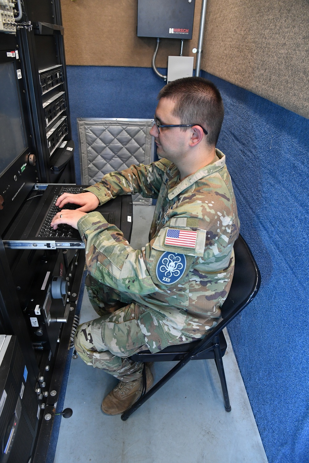 A U.S. military service member in OCP (Operational Camouflage Pattern) uniform is seated at a rack-mounted computer workstation, typing on a keyboard. The individual wears a U.S. flag patch and a blue circular unit patch featuring an octopus and Roman numeral 'XXII' on the right sleeve. The setting appears to be a secure or specialized technical operations room with server rack equipment.