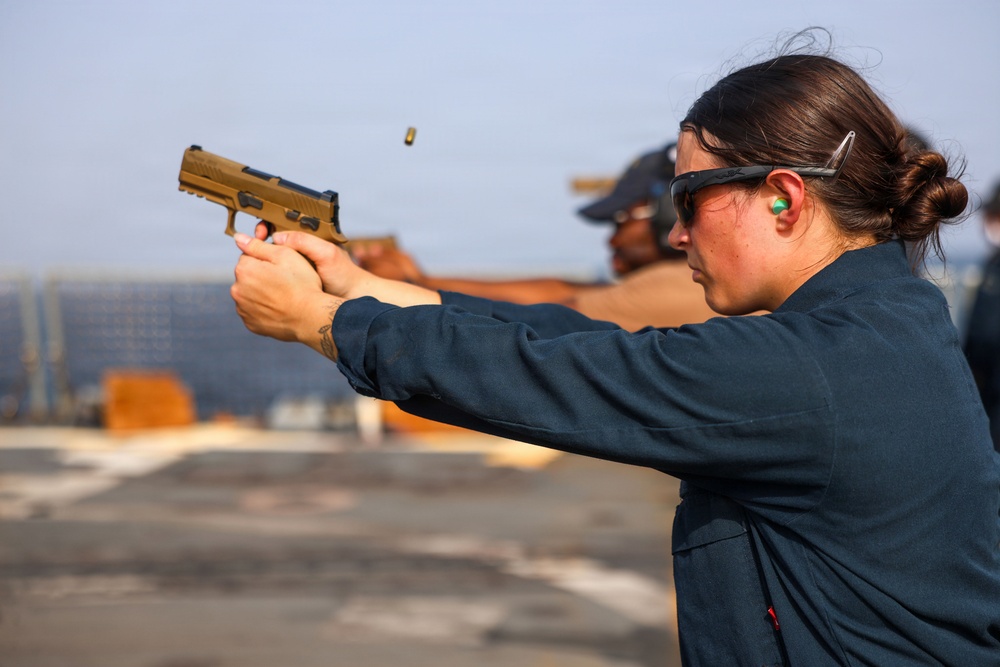 A person wearing protective eyewear and ear protection is firing a tan/FDE-colored semi-automatic pistol at what appears to be an outdoor shooting range. A spent brass casing is visibly ejecting from the firearm mid-shot. The setting appears to be on a flat, open surface, possibly a military vessel or rooftop range, with another individual visible in the background.