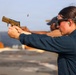 A person wearing protective eyewear and ear protection is firing a tan/FDE-colored semi-automatic pistol at what appears to be an outdoor shooting range. A spent brass casing is visibly ejecting from the firearm mid-shot. The setting appears to be on a flat, open surface, possibly a military vessel or rooftop range, with another individual visible in the background.