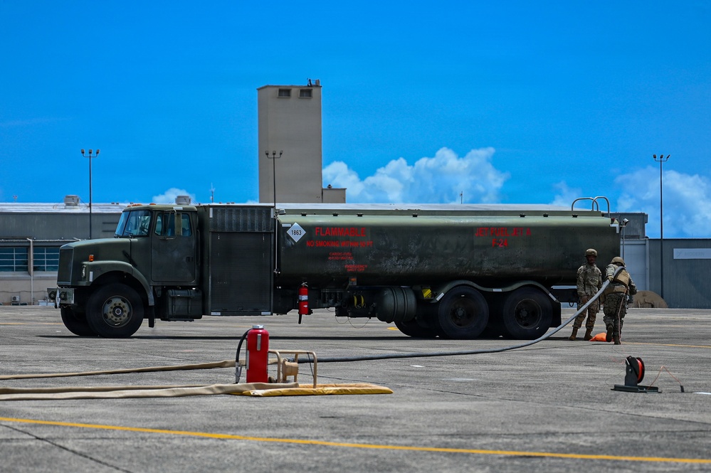 A military fuel tanker truck labeled 'FLAMMABLE' and 'JET FUEL JP-8' is parked on an airfield tarmac. Two personnel in military uniforms are working near the rear of the vehicle, managing fuel hoses. The scene appears to be a routine aircraft refueling operation at a military air base.