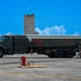 A military fuel tanker truck labeled 'FLAMMABLE' and 'JET FUEL JP-8' is parked on an airfield tarmac. Two personnel in military uniforms are working near the rear of the vehicle, managing fuel hoses. The scene appears to be a routine aircraft refueling operation at a military air base.