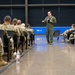 A military briefing or training session is taking place in a modern indoor facility with a polished concrete floor. A single presenter wearing an olive green flight suit is addressing a large group of soldiers seated in rows on both sides, all dressed in camouflage uniforms. The presenter is holding a notebook or binder and appears to be speaking to the assembled personnel.