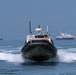 A military or law enforcement rigid inflatable boat (RIB) is moving at speed toward the camera, creating a prominent wake. Personnel in military/tactical gear are visible aboard the vessel, which is equipped with navigation and communication equipment. Several larger vessels are anchored or moored in the background harbor.