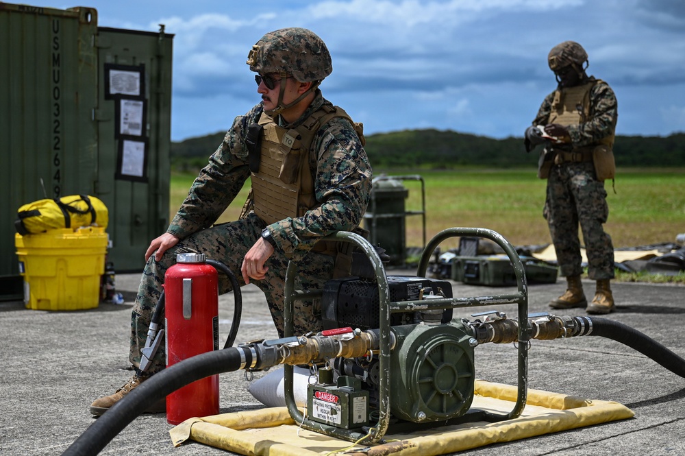 Two military personnel in MARPAT camouflage uniforms and tactical gear are conducting what appears to be a fuel handling or fluid transfer operation at an outdoor military installation. The foreground soldier is positioned next to a fuel pump unit with hoses attached, alongside a red fire extinguisher as a safety precaution. A second soldier stands in the background appearing to review documentation or a device.