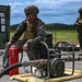 Two military personnel in MARPAT camouflage uniforms and tactical gear are conducting what appears to be a fuel handling or fluid transfer operation at an outdoor military installation. The foreground soldier is positioned next to a fuel pump unit with hoses attached, alongside a red fire extinguisher as a safety precaution. A second soldier stands in the background appearing to review documentation or a device.