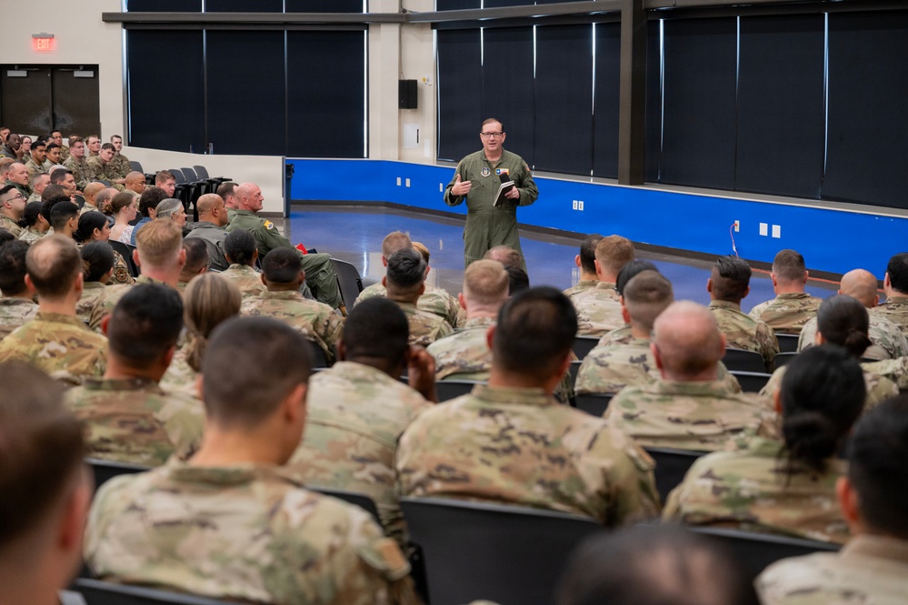 A military officer in a green flight suit addresses a large assembly of uniformed service members seated in an auditorium or briefing hall. The speaker appears to be holding a small notebook or document while delivering remarks to the audience. The majority of attendees are wearing OCP (Operational Camouflage Pattern) uniforms, suggesting a mixed-branch or Air Force unit gathering.