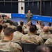 A military officer in a green flight suit addresses a large assembly of uniformed service members seated in an auditorium or briefing hall. The speaker appears to be holding a small notebook or document while delivering remarks to the audience. The majority of attendees are wearing OCP (Operational Camouflage Pattern) uniforms, suggesting a mixed-branch or Air Force unit gathering.