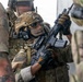The image shows multiple individuals in full tactical/military gear in what appears to be a training or operational environment. The central figure is holding a rifle with an optic and tactical light, wearing a ballistic helmet, ear protection, sunglasses, and a face covering. The scene depicts a close-quarters tactical scenario with multiple armed personnel in multicam camouflage.