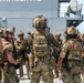 A group of heavily equipped tactical personnel in multicam camouflage uniforms are gathered on what appears to be the deck of a naval vessel. The central figure's backpack is clearly labeled 'United States Coast Guard,' and several personnel display flag patches consistent with the United States and Peru, indicating a joint multinational operation. The group appears to be conducting a briefing or coordination meeting near the ship's hull.
