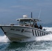 A military/law enforcement patrol boat designated BPC-3707, belonging to the 'Aeronaval' unit, is underway at speed on open water. The vessel carries multiple armed personnel in military gear and helmets. Several large cargo ships are visible in the background anchorage.