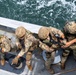 Four military personnel in full combat gear are positioned along the edge of a naval vessel deck, overlooking open water. They are wearing multicam camouflage uniforms and tactical helmets, with at least one operator visibly holding an assault rifle. The overhead angle suggests this is a maritime boarding or vessel interdiction training exercise.