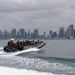 A military rigid inflatable boat (RIB) carrying multiple armed personnel in tactical gear is moving at high speed across open water. In the background, a dense urban skyline with numerous high-rise buildings is visible under overcast skies. The scene suggests a military or law enforcement maritime operation near a major coastal city.