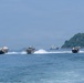 Three fast-moving patrol or interceptor boats are traveling at high speed across open water, leaving prominent white wake trails behind them. In the background, several larger vessels including what appears to be a cruise ship and fishing/cargo boats are anchored or moving slowly near a lush, tree-covered island. The scene suggests a maritime patrol or escort operation in a coastal bay area.
