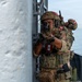 Two military personnel in full tactical gear are conducting what appears to be a maritime boarding or ship-clearing exercise on the deck of a naval vessel. Both operators display Peruvian flag patches on their helmets and uniforms, identifying them as members of the Peruvian armed forces. The lead operator is aiming a firearm around a corner while the second provides rear security.