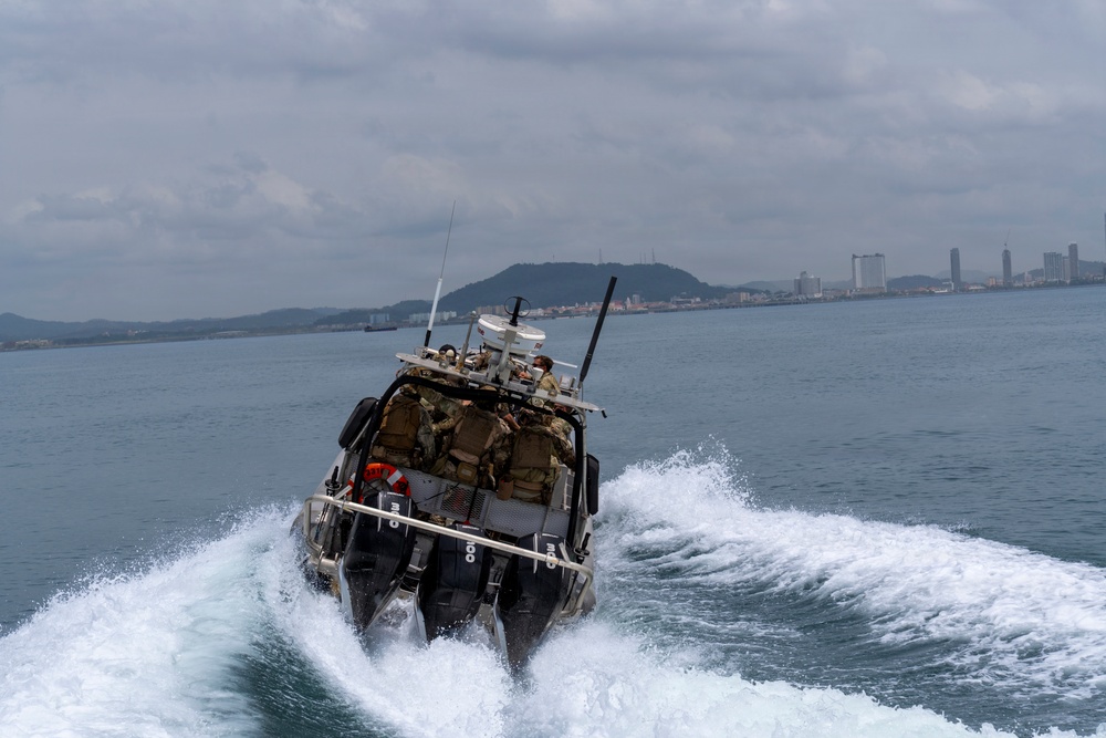 A military or law enforcement tactical boat is moving at high speed across open water, creating a large white wake. Several personnel in military-style tactical gear are visible aboard the vessel, which is equipped with communications antennas and surveillance equipment. A coastal city skyline and green hills are visible in the background under overcast skies.