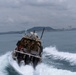 A military or law enforcement tactical boat is moving at high speed across open water, creating a large white wake. Several personnel in military-style tactical gear are visible aboard the vessel, which is equipped with communications antennas and surveillance equipment. A coastal city skyline and green hills are visible in the background under overcast skies.