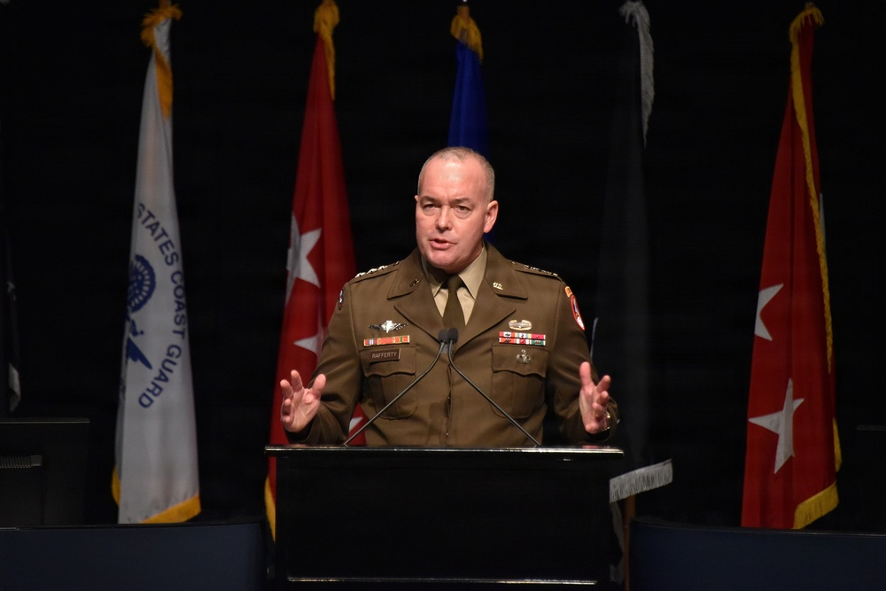 A military officer in an olive/tan dress uniform is speaking at a podium during a formal military ceremony or conference. The officer's name tag reads 'RAFFERTY' and they are gesturing with both hands while addressing an audience. Multiple military service flags are displayed in the background.
