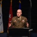 A military officer in an olive/tan dress uniform is speaking at a podium during a formal military ceremony or conference. The officer's name tag reads 'RAFFERTY' and they are gesturing with both hands while addressing an audience. Multiple military service flags are displayed in the background.