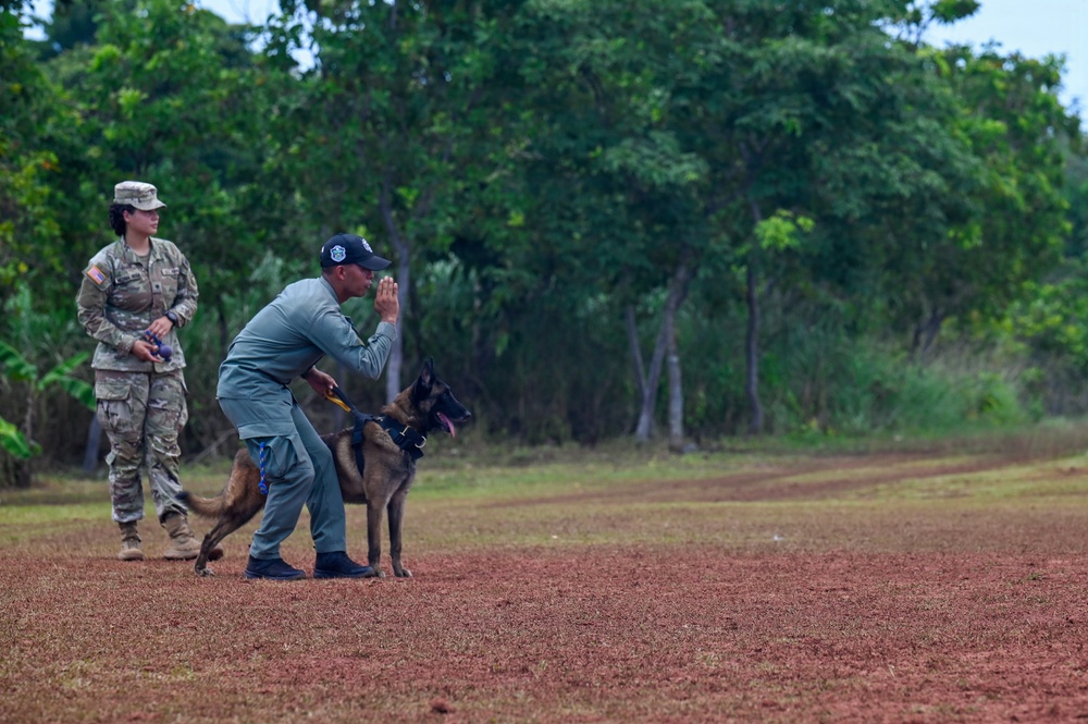 A dog handler in gray uniform and dark cap is giving a hand signal command to a Belgian Malinois on a red dirt field. A U.S. Army soldier in full camouflage uniform with American flag patch observes the demonstration from behind. The setting appears to be a military working dog training exercise conducted outdoors near a tree line.