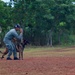 A dog handler in gray uniform and dark cap is giving a hand signal command to a Belgian Malinois on a red dirt field. A U.S. Army soldier in full camouflage uniform with American flag patch observes the demonstration from behind. The setting appears to be a military working dog training exercise conducted outdoors near a tree line.