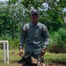A uniformed law enforcement officer stands outdoors with a Belgian Malinois K-9 on a leash and harness. The officer wears a gray-green uniform with a Panamanian flag patch on the left shoulder, a name tape reading 'L. FERNANDEZ,' and a cap with an insignia. The setting appears to be a training or outdoor operational area surrounded by tropical vegetation.
