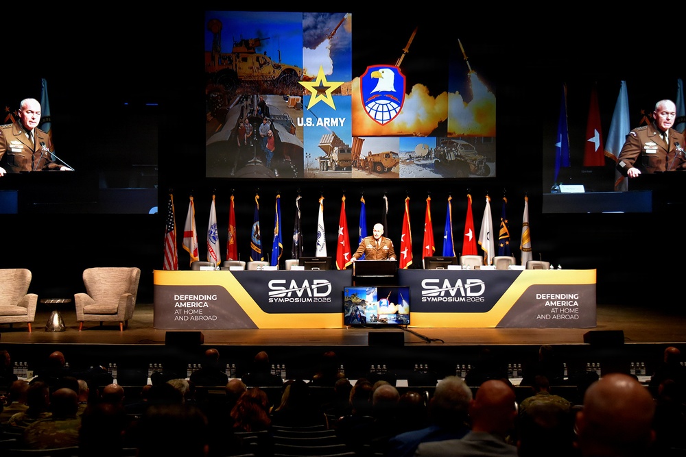 A military officer in brown uniform delivers a keynote address at the SMD Symposium 2026, a U.S. Army Space and Missile Defense conference. The stage features a large backdrop displaying U.S. Army and SMDC (Space and Missile Defense Command) insignia alongside imagery of military equipment. The event theme is 'Defending America at Home and Abroad,' with a large audience of military and civilian personnel in attendance.