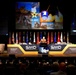 A military officer in brown uniform delivers a keynote address at the SMD Symposium 2026, a U.S. Army Space and Missile Defense conference. The stage features a large backdrop displaying U.S. Army and SMDC (Space and Missile Defense Command) insignia alongside imagery of military equipment. The event theme is 'Defending America at Home and Abroad,' with a large audience of military and civilian personnel in attendance.