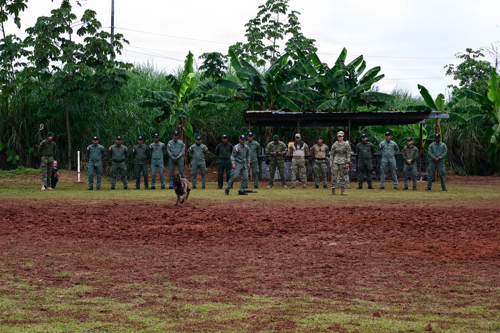 A group of approximately 20 uniformed personnel are lined up along the edge of a red dirt training field in a tropical setting. A military working dog is visible in the foreground of the field, suggesting an active K9 demonstration or training exercise. Two individuals in camouflage uniforms distinct from the majority appear to be advisors or instructors observing alongside the green-uniformed group.