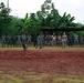 A group of approximately 20 uniformed personnel are lined up along the edge of a red dirt training field in a tropical setting. A military working dog is visible in the foreground of the field, suggesting an active K9 demonstration or training exercise. Two individuals in camouflage uniforms distinct from the majority appear to be advisors or instructors observing alongside the green-uniformed group.