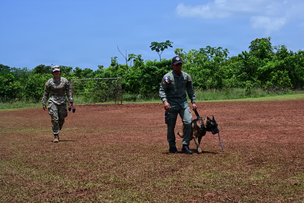 Two uniformed personnel are walking across an open red-dirt field with lush green vegetation in the background. One individual wearing US military camouflage (MultiCam/OCP pattern) walks behind another person in olive/gray uniform bearing what appears to be a Panamanian flag patch, who is handling a Belgian Malinois on a leash. The setting and uniforms suggest a joint military or law enforcement K9 training exercise.