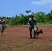 Two uniformed personnel are walking across an open red-dirt field with lush green vegetation in the background. One individual wearing US military camouflage (MultiCam/OCP pattern) walks behind another person in olive/gray uniform bearing what appears to be a Panamanian flag patch, who is handling a Belgian Malinois on a leash. The setting and uniforms suggest a joint military or law enforcement K9 training exercise.