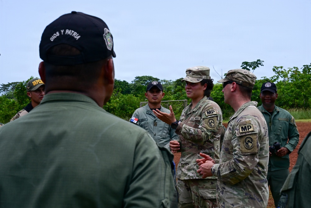 The image shows a group of U.S. Army Military Police (MP) soldiers engaged in a discussion with what appear to be Panamanian security forces personnel, likely during a joint training or liaison event. Two U.S. Army soldiers wearing MultiCam uniforms with 'MP' patches and Africa Command shoulder sleeve insignia are speaking with personnel in olive-green uniforms. One individual in the foreground wears a cap reading 'Dios y Patria' (God and Homeland), a Panamanian national motto, suggesting a bilateral military cooperation context.
