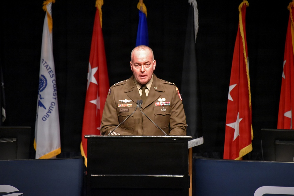 A U.S. Army officer identified by nameplate as 'RAFFERTY' is speaking at a podium during what appears to be an official military ceremony or conference. He is wearing a Class A Army Service Uniform with multiple medals, badges, and rank insignia. Multiple military service flags are displayed behind him, including a visible U.S. Coast Guard flag.