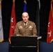 A U.S. Army officer identified by nameplate as 'RAFFERTY' is speaking at a podium during what appears to be an official military ceremony or conference. He is wearing a Class A Army Service Uniform with multiple medals, badges, and rank insignia. Multiple military service flags are displayed behind him, including a visible U.S. Coast Guard flag.