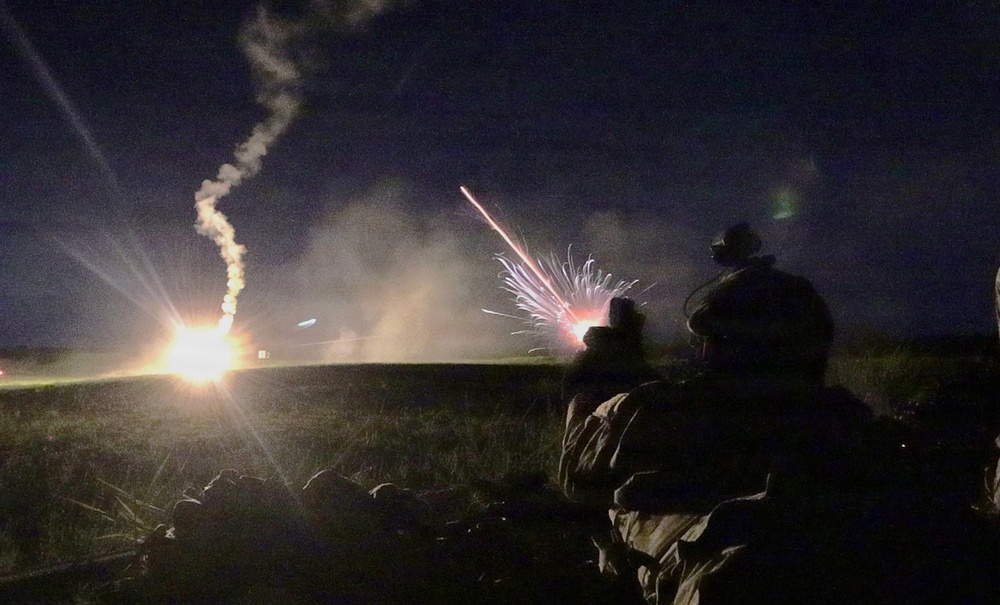 A nighttime military training or combat exercise showing soldiers in the foreground observing multiple pyrotechnic or weapons discharges in the distance. At least two distinct light events are visible: a bright ground-level explosion or flare on the left and a starburst/white phosphorus-type burst in the center. The scene is illuminated primarily by the weapons fire against a dark sky.